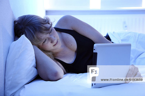 Junge Frau im Bett surft mit einem iPad Tablet-Computer per Drahtlosverbindung im Internet