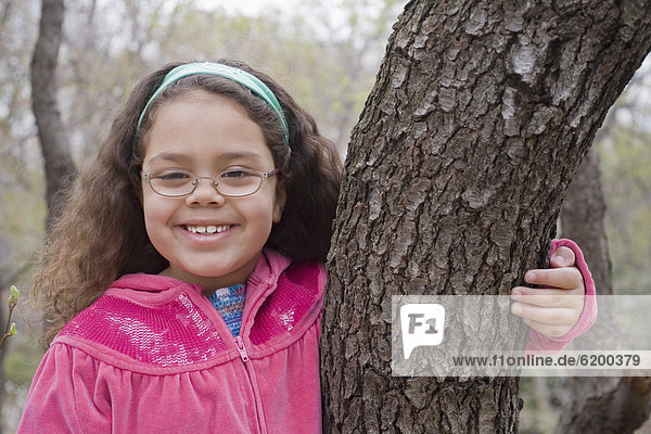Smiling Hispanic girl hugging tree trunk