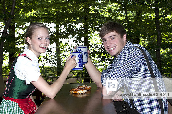 Junges Paar mit Dirndl und Lederhose stößt mit Bierkrügen an  im Biergarten  bei Pettstatt  Oberfranken  Bayern  Deutschland  Europa