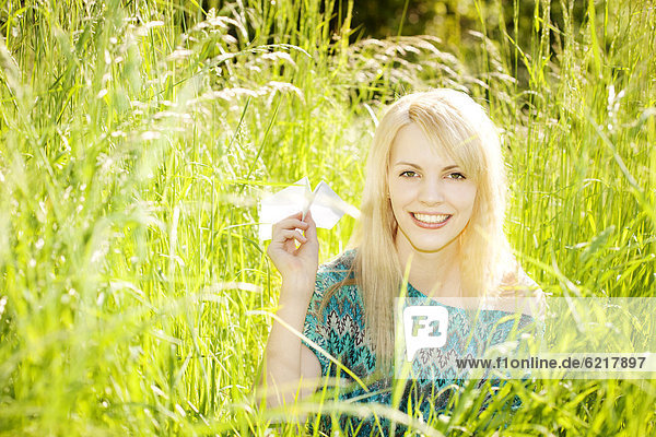 Smiling young woman sitting in long grass  throwing paper plane