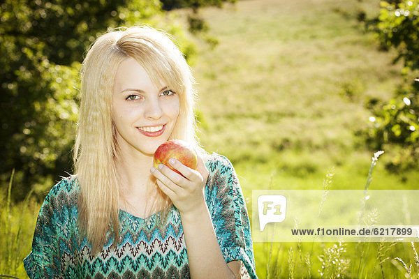 Lächelnde junge Frau sitzt lachend mit einem Apfel auf einer Wiese