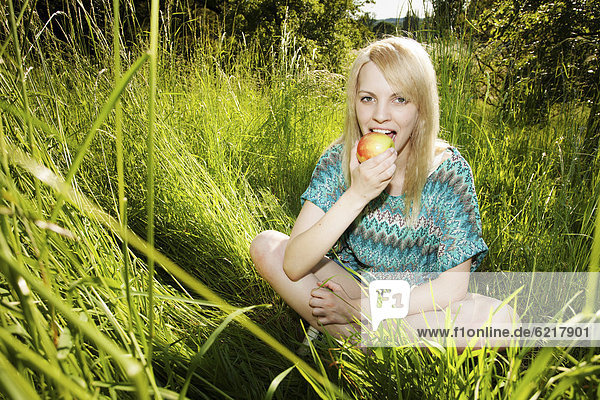 Smiling young woman sitting in tall grass eating an apple