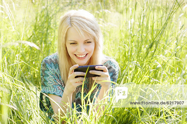 Junge Frau sitzt auf einer Wiese im hohen Gras und kommuniziert per Mobiltelefon