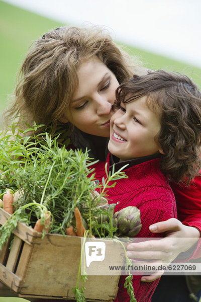 Boy holding a crate of vegetables with his mother kissing him