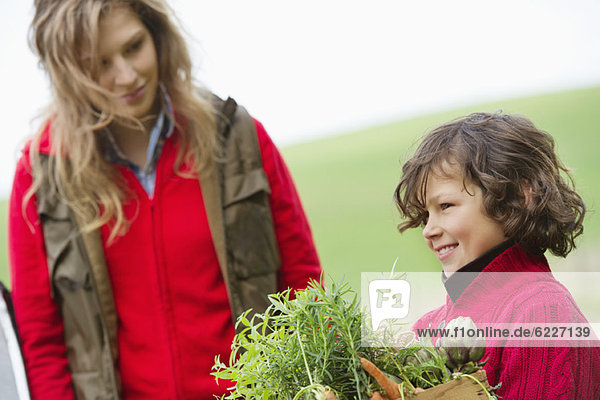 Junge hält eine Kiste mit verschiedenen Gemüsesorten mit seiner Mutter auf einem Bauernhof.