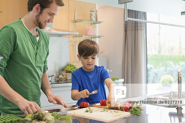 Mann schaut seinen Sohn an  wie er in der Küche Gemüse schneidet.