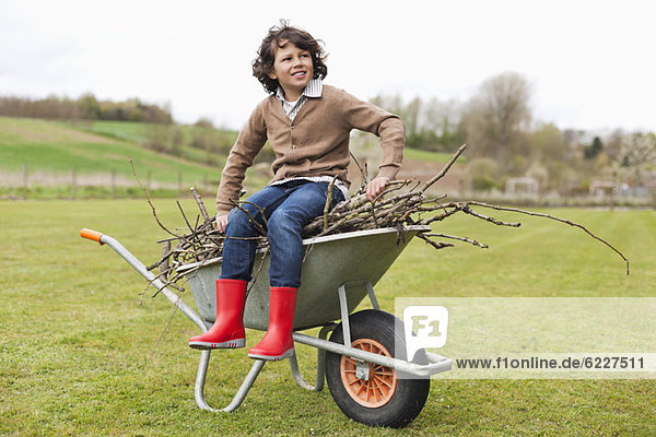 Boy sitting on a wheelbarrow with firewood in a field