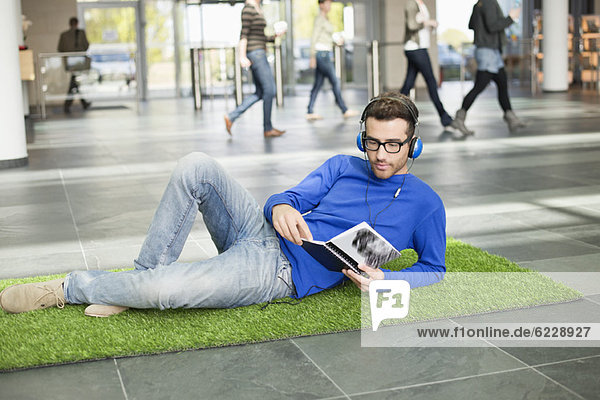 Geschäftsmann hört Musik und liest Buch  während er sich auf einer Grasmatte in einer Bürolobby ausruht.