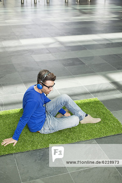 Geschäftsmann entspannt sich auf Rasen und hört Musik in einer Büro-Lobby.