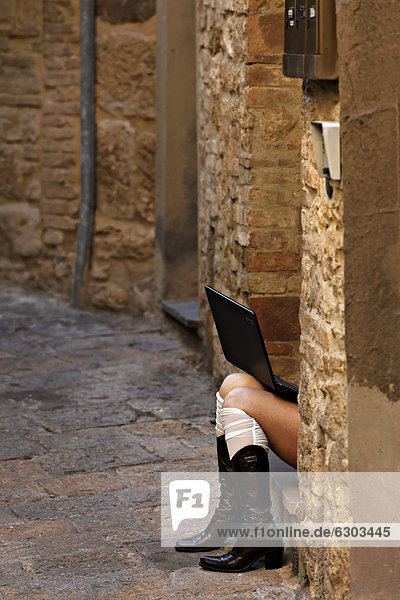 Legs of a woman sitting in doorway wearing fashion boots and using a laptop  Volterra  Tuscany  Italy  Europe