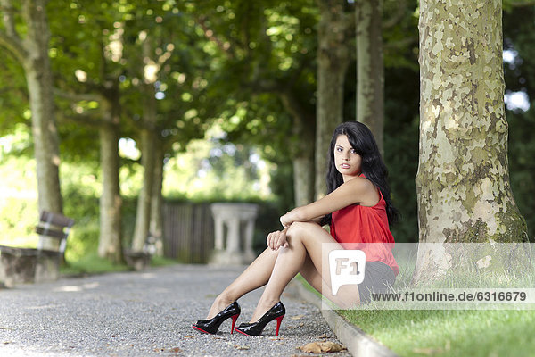 Young woman in a red top  black hot pants and high heels sitting down in front of a tree