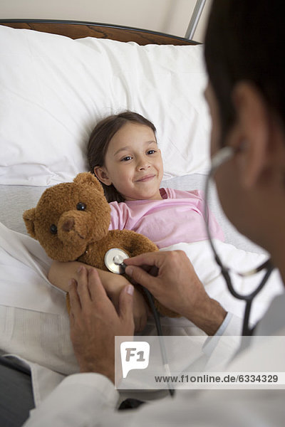 Arzt tröstet junge Patientin  indem er ihren Teddybär mit einem Stethoskop untersucht.