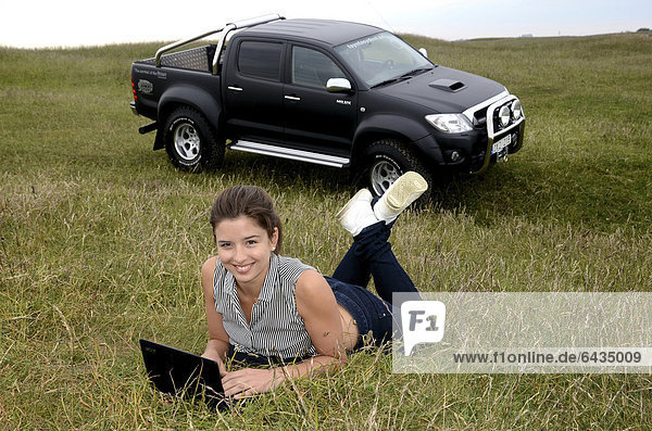Junge Frau mit Laptop-Computer vor einem Toyota Hilux im Gras liegend  Schweden  Europa