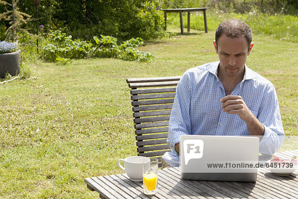 A man sitting at a table in his backyard having breakfast and using a laptop