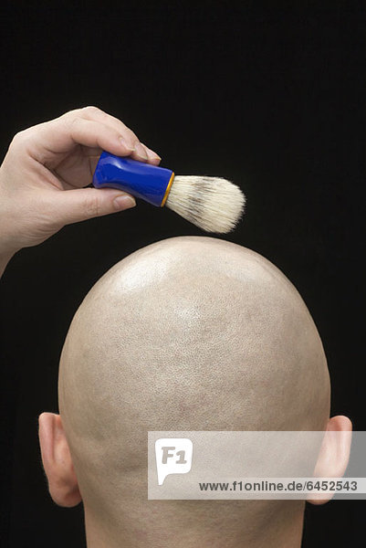 Eine Frau hält einen Rasierpinsel über dem rasierten Kopf eines Mannes.