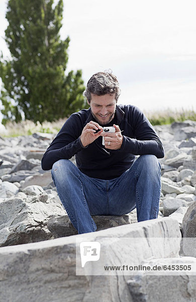 Ein Mann sitzt auf einem Felsen und lächelt etwas auf seinem Smartphone an.