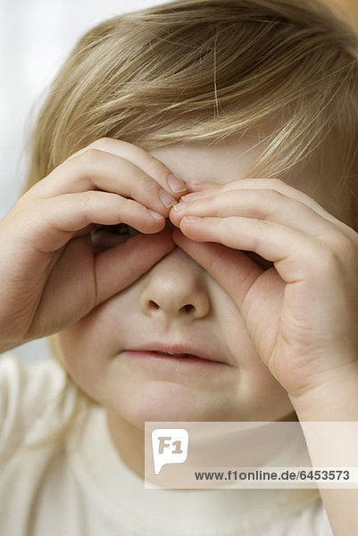 Ein kleines Mädchen schaut durch eine Brille aus ihren Fingern.