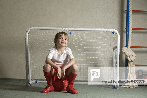 Ein Junge sitzt auf einem Fußball vor einem Fußballtor.