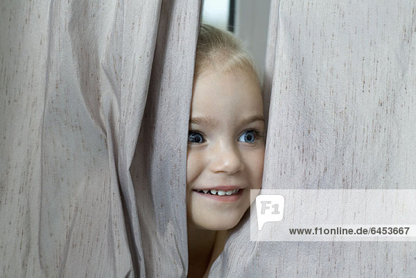 Ein junges Mädchen  das sich zwischen Fenstervorhängen versteckt.