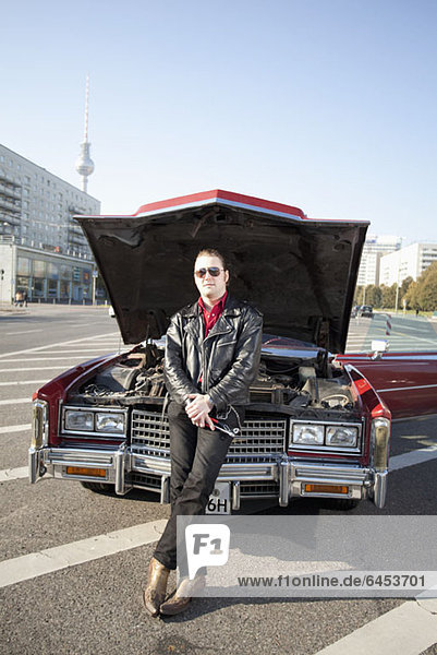 A cool rockabilly guy holding tools leaning against the front of his vintage car  hood up