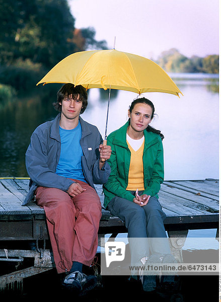 1216669 outdoor lake water pier day spring couple girl woman brunette blouse green trousers orange grey jacket young man boy 20-25 dark haired sit hold yellow umbrella vacation holidays rest relax vertical