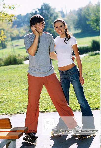 1216721 outdoor park day spring couple girl woman brunette long hair grey t-shirt t shirt white blouse jean jeans trousers orange young man boy 20-25 dark haired roller skates skate stand sport recreation smile smiling hold telephone phone cell cellular cellphone mobile call talk speak communication telecommunication vertical