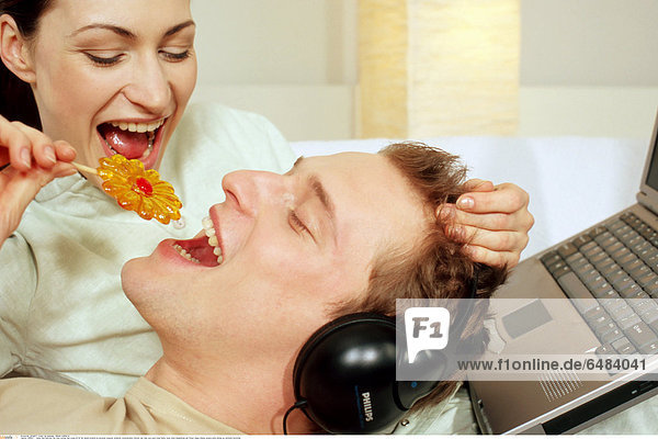 1218821 indoor bed bedroom flat room woman man young 25-30 fair haired brunette lie personal computer notebook communication internet rest relax eye eyes close hobby music listen headphones eat flower shape lollipop sweets smile smiling joy emotions horizontal