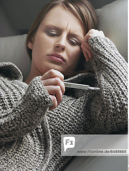 1221079 indoor flat room young woman 25-30 brunette grey sweater lie sofa couch hold thermometer fever temperature look read ill disease illness cold resist hand close up vertical