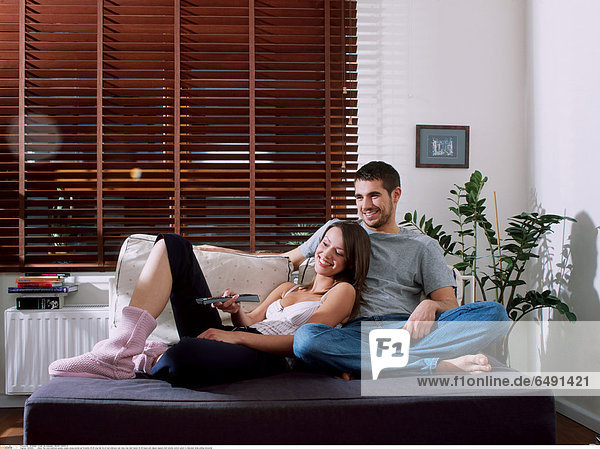 1234433 indoor flat room bedroom people couple young woman girl brunette 20-25 long hair lie sit bed embrace rest relax man dark haired 25-30 beard pink slipper slippers hold remote control watch tv television smile smiling horizontal