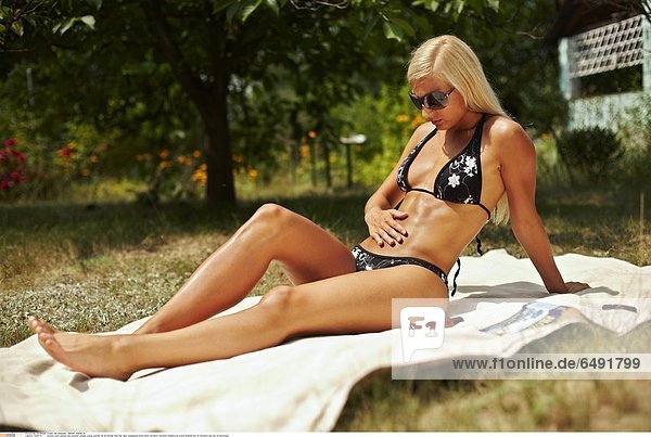 1235775 outdoor park garden day summer people young woman 25-30 blonde long hair dark sunglasses body bikini swimsuit vacation holidays sit towel sunbath put on stomach sun tan oil horizontal