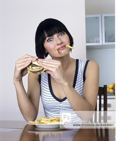 1239119 indoor flat kitchen people woman young girl 20-25 brunette fringe blouse white stripe stripes striped eat plate hamburger fast food chip chips vertical smile smiling close up portrait mime fun play