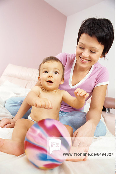 1239166 indoor flat room people boy girl child 0-5 baby blonde fair haired white sheet sit stand mother motherhood smile smiling naked nakedness woman young 25-30 brunette blouse pink vertical ball hold play fun embrace
