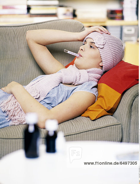 1245202 indoor flat room people young woman 20-25 girl lie sofa couch cap scarf ill illness cold bottle syrup pour drug medicine measure take temperature thermometer fever close up vertical