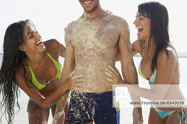 Two women rubbing sand on man at beach