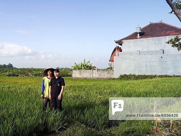 Ngurah  a young rice farmer stands in a rice paddy next to his wife who is dressed to go to work at a popular restaurant and club for foreigners In the background  villas have been built on the rice paddy  showing some of the breakdown of the 3 vital pil. Ngurah  a young rice farmer stands in a rice paddy next to his wife who is dressed to go to work at a popular restaurant and club for foreigners In the background  villas have been built on the rice paddy  showing some of the breakdown of the 3 vital pillars of Subak culture - land  water and labor The Subak is Klepekan in Bali