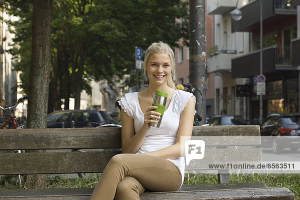 Germany  North Rhine Westphalia  Cologne  Young woman sitting on bench with coffee cup  smiling  portrait