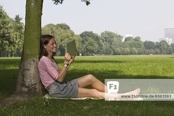 Germany  North Rhine Westphalia  Cologne  Young student sitting in park with digital tablet  smiling