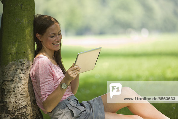 Deutschland  Nordrhein-Westfalen  Köln  Junge Studentin im Park mit digitalem Tablett sitzend  lächelnd