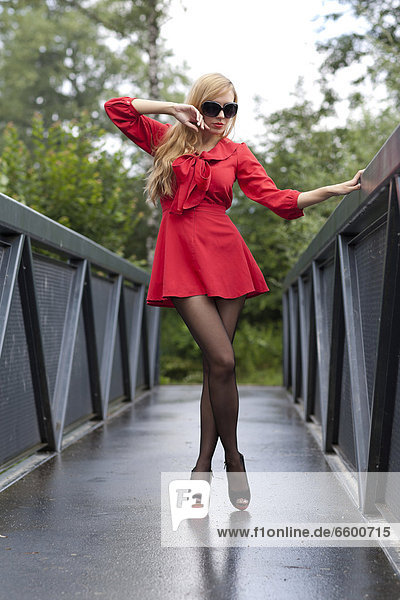 Young woman with sunglasses in a short red dress and high heels posing on a bridge