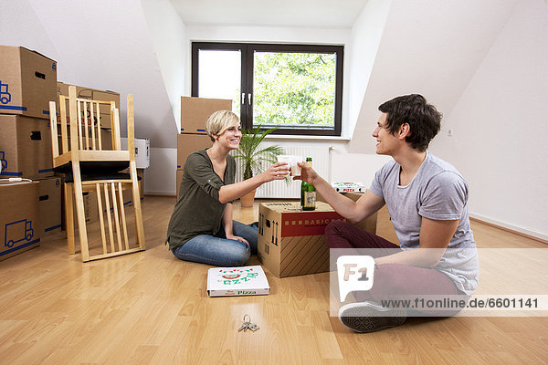 Young couple sitting in an empty room in a new apartment and raising their glasses of champagne to celebrate moving in together
