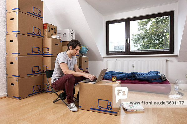 Young man sitting in an empty room in a new apartment  provisionally furnished with a sleeping bag  an air mattress  a laptop and moving boxes