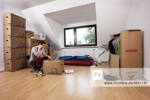 Young man sitting in an empty room in a new apartment  provisionally furnished with a sleeping bag  an air mattress  a laptop and moving boxes
