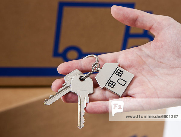Hand holding house keys with a metal key tag shaped like a house  moving boxes at the back