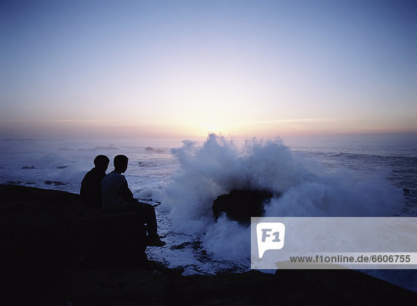 Two Young Boys Watching Waves Coming Crashing Into Rocks And The Sea Walls Of Essaouira At Dusk