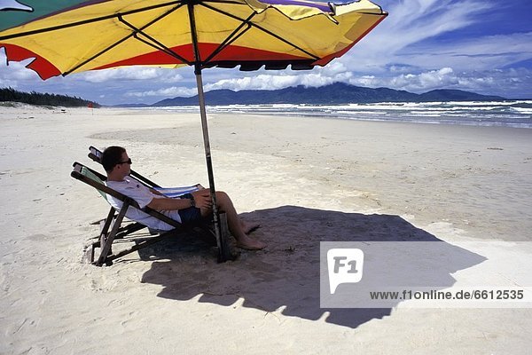 sitzend Mann Strand Regenschirm Schirm unterhalb Liege Liegen Liegestuhl Seitenansicht Sonnenschirm Schirm