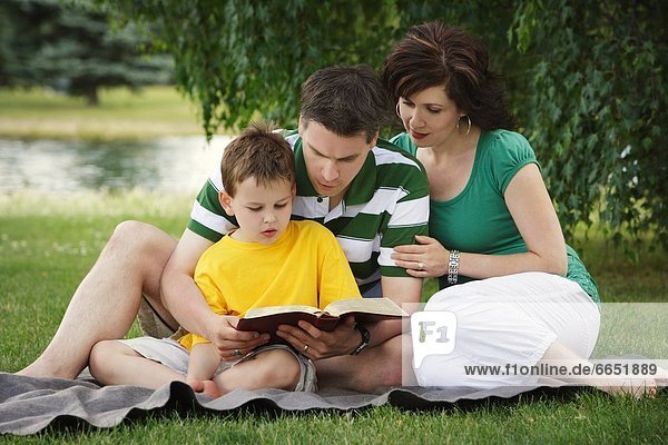 Family Sitting On Blanket In Park And Studying The Bible Together