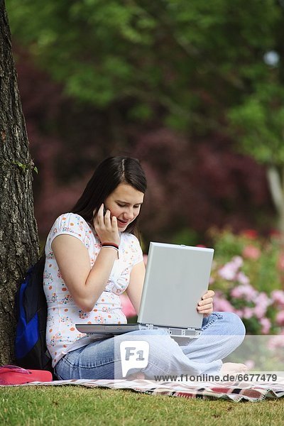 A Teenage Girl Working On Her Laptop Computer And Talking On Her Cellphone Under A Tree