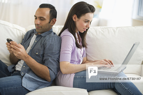 Couple sitting on sofa back to back using laptop and phone