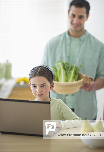 Girl (8-9) using laptop and father preparing food in domestic kitchen