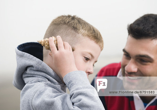 Father watching son hold seashell up to ear
